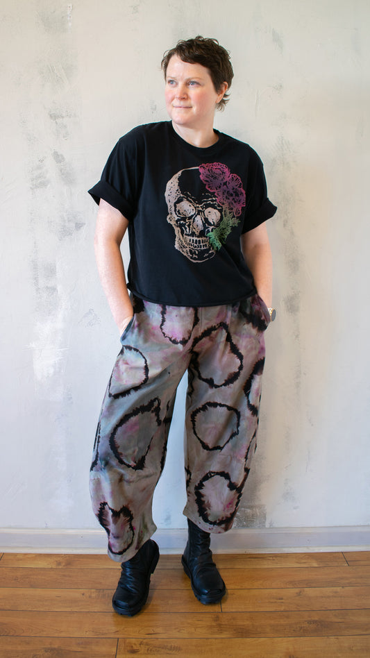 Person wearing a black t-shirt with a colorful skull design and patterned pants against a plain wall.